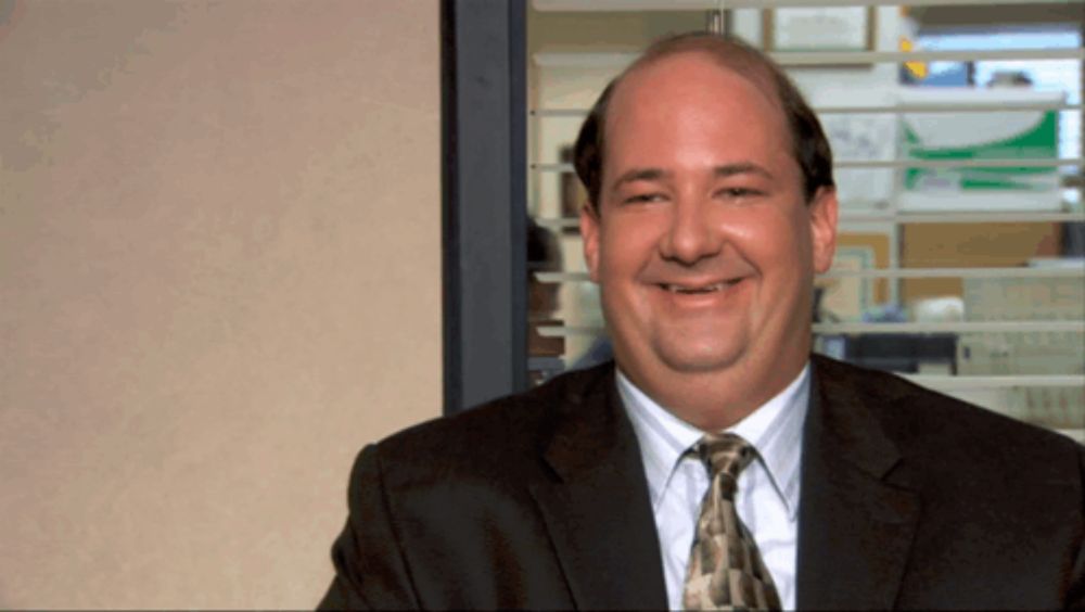 a bald man in a suit and tie smiles in front of a window