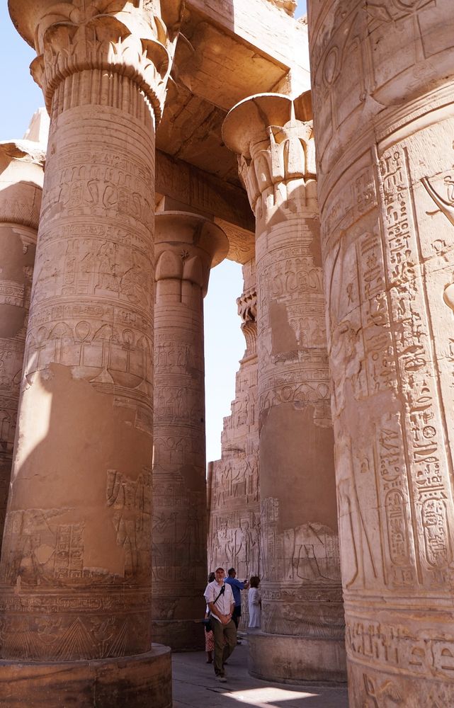 Photograph showing people looking very small next to massive stone pillars at a temple in Egypt. The pillars have carvings of hieroglyphics over most of them, although in some areas time or damage has worn the stone smooth. 