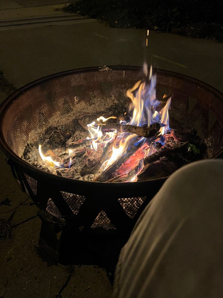 Fire pit with flames in the dark, gray knee of jeans in the foreground. 