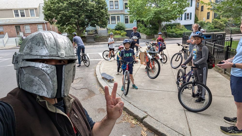 a guy in a Mandolarian helmet from Star Wars with a bunch of kids getting ready to bike