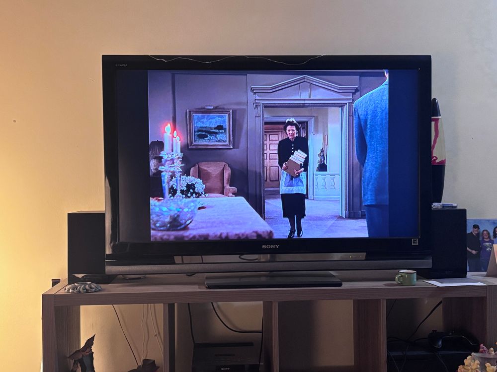 A tv showing a shot of a maid in the middle of the shot carrying a stack of books getting closer to a table with candle holders and lit candles on it in the foreground.