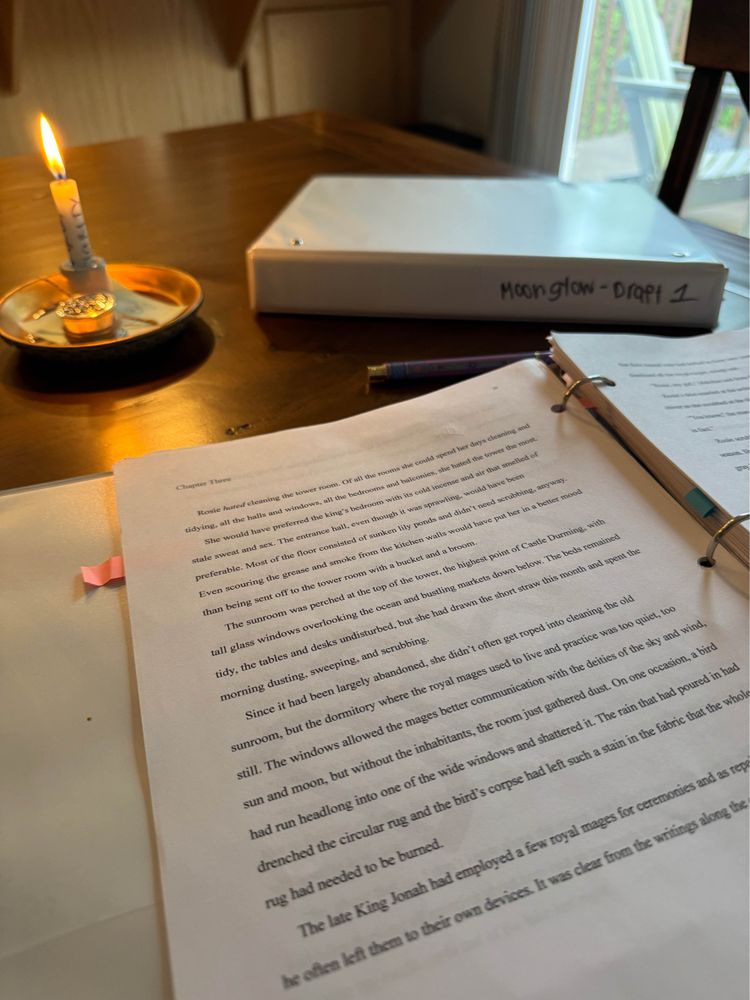 Two binders, one open with a typed document, the other marked MOONGLOW - DRAFT 1. A small candle burning to one side. 