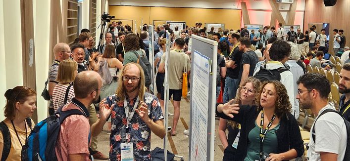 The image captures a vibrant poster session at the useR! R Conference, held in Salzburg in July 2024. The setting is a spacious conference hall with attendees engaging in discussions around research posters mounted on large display boards. Participants, a mix of data scientists, researchers, and R enthusiasts, are seen standing in small groups, gesturing towards the posters, and exchanging insights. Some individuals are holding conference materials or notebooks, while others are listening attentively to presenters explaining their work. The posters feature graphs, charts, and R code snippets, reflecting cutting-edge developments in R programming for statistical analysis and data science. The atmosphere is collaborative and professional, with attendees dressed in business casual attire. In the background, additional posters and conference branding related to R programming and statistical computing reinforce the theme of the event. The lighting in the room is bright, enhancing visibility and interaction.

