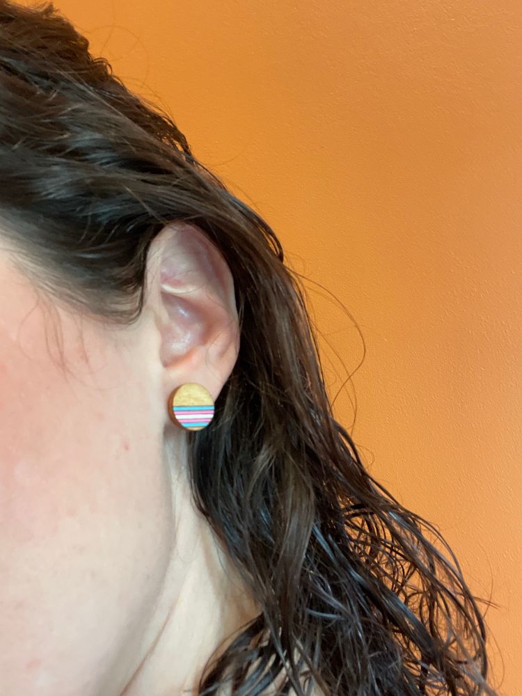 Close shot of a woman’s ear with earring. The earring is a small circle of natural wood, with the lower half of it carved in horizontal lines. The lines are painted the trans pride flag colors of (top to bottom) blue, pink, white, pink, and blue. The woman has dark, curly hair, pale skin, and is against a bright orange wall. 