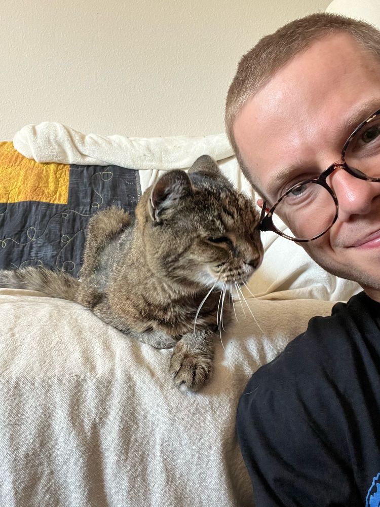 On the right, a man sitting in front of a couch smiles at the camera. In the center, a happy tabby cat rubs her head on the corner of his glasses.