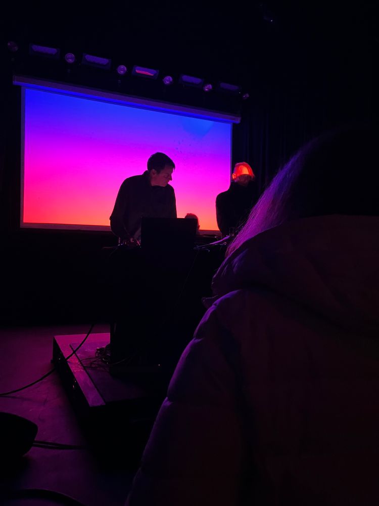 Ambient musicians Loscil and Lawrence English performing in front of a screen of blue, pink, and orange