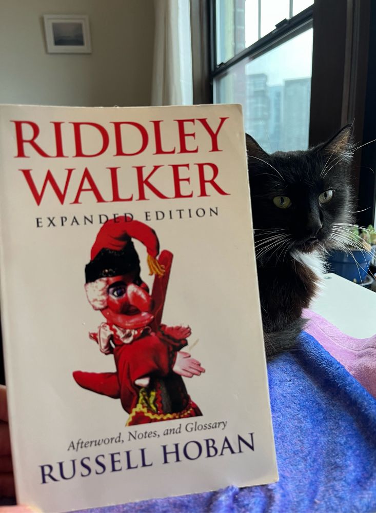 The cover of Russell Hoban’s novel “Riddley Walker,” showing a puppet against a white background. A cat looks at the camera from behind the book.