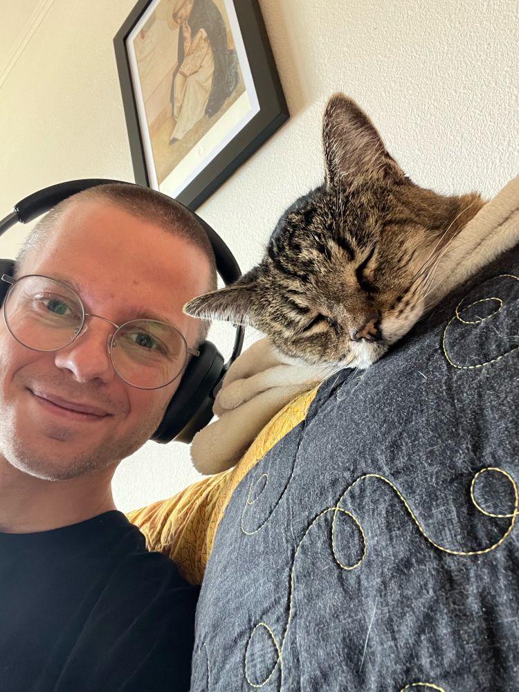 On the left, a man wearing headphones and glasses smiles at the camera sitting on a couch. On the right, above his shoulder, a beautiful gray tabby cat sleeps facing the camera on the back of three couch.