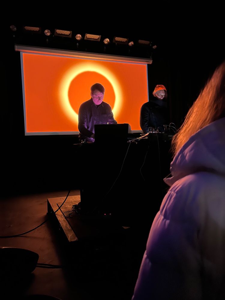 Ambient musicians Loscil and Lawrence English performing in front of a deep orange screen