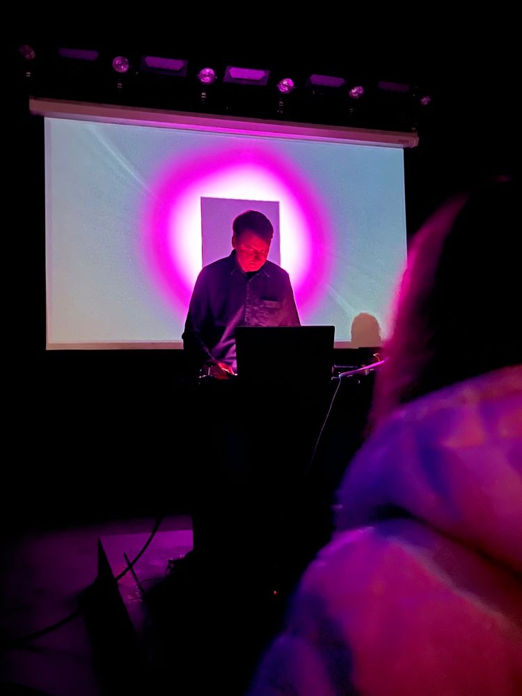 Ambient musicians Loscil and Lawrence English performing in front of a screen of grey, pink, and white