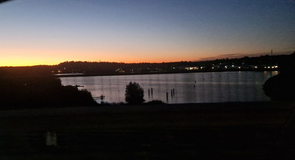 Morgenstimmung mit Blick auf einen See. Am Horizont färbt die aufgehende Sonne den Himmel orange, während er nach oben in dunkles Blau übergeht. Silhouetten von Bäumen und Gebäuden zeichnen sich im Vordergrund und am Ufer ab. Auf der rechten Seite sind viele kleine Lichter einer Stadt die sich im Wasser spiegeln.