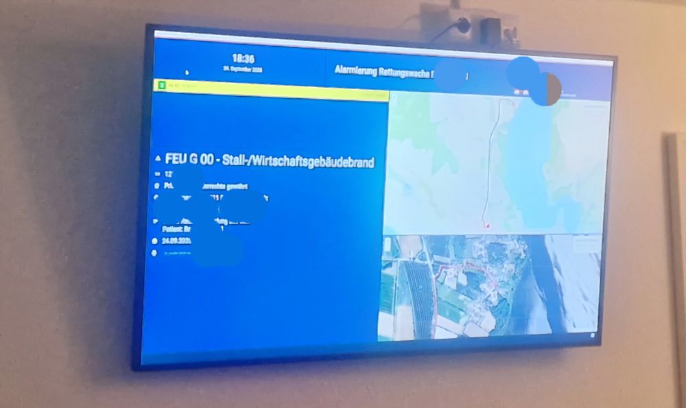 Ein Wandbildschirm zeigt eine Einsatzübersicht der Leitstelle. Auf blauem Hintergrund steht der Einsatztext: „FEU G 00 – Stall-/Wirtschaftsgebäudebrand“. Rechts sind Kartenansichten mit einer markierten Route und einem Luftbild der Umgebung eingeblendet. Oben im Bildschirm ist die Uhrzeit 18:36 sowie das Datum 24. September 2025 sichtbar. Teile sensibler Daten sind unkenntlich gemacht.
