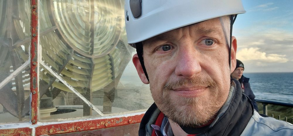 Nahaufnahme einer Person mit weißem Schutzhelm und Jacke, die vor der großen Glaslinse eines Leuchtturms steht. Im Hintergrund sind Meer, Himmel mit Wolken und eine weitere Person mit Mütze erkennbar.