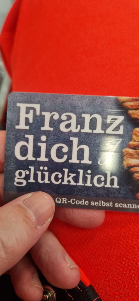 Eine Hand hält eine rechteckige Werbekarte. Darauf steht in großer weißer Schrift: „Franz dich glücklich“. Im Hintergrund ist eine dunkle, strukturierte Fläche, am rechten Rand ist ein Stück eines Gebäcks (vermutlich Franzbrötchen) sichtbar. Unten am Rand steht in kleinerer Schrift: „QR-Code selbst scannen“.