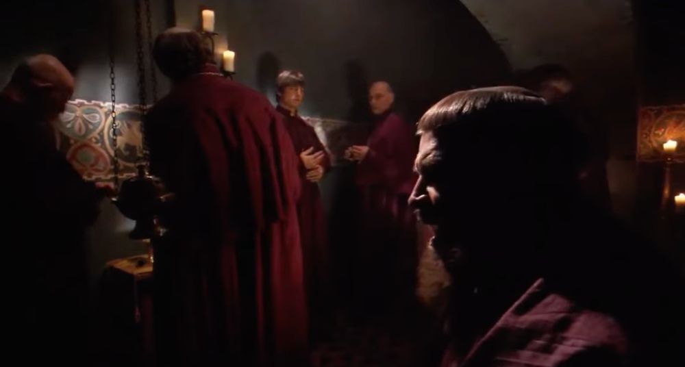 Brian Blessed as Piccolomini in profile, seated on the toilet while other cardinals react with embarrassment and disgust.