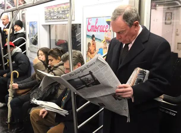 Michael Bloomberg in the NYC subway, standing at a door reading the NYTimes.