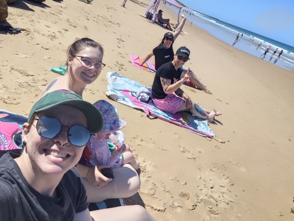 Four adults and a child on a sunny beach, with yellow sand and blue water. They are all sitting on multicoloured beach towels, and looking at the camera while smiling. The photo is being taken as a selfie. 