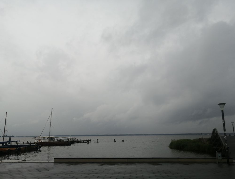 Graue Wolken, graues Wasser. Links dunkle Boote. Rechts ein Steg. 
