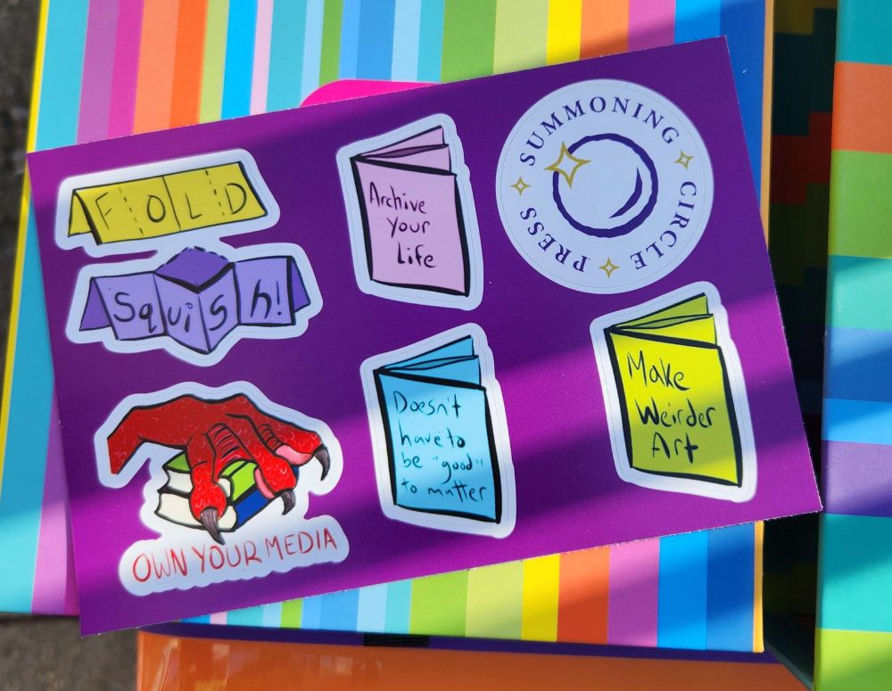 A sticker sheet of drawn illustrations about zines and a claw over books and the Summoning Circle Press logo. The printed sheet sits on a colourful box.