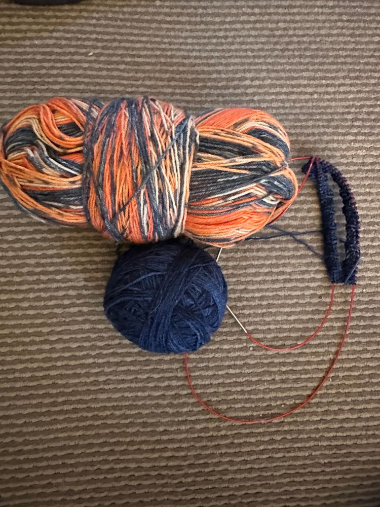 Skein of orange, blue and cream yarn above a ball of navy yarn next to the top of the cuff of a navy sock on a long circular knitting needle. 