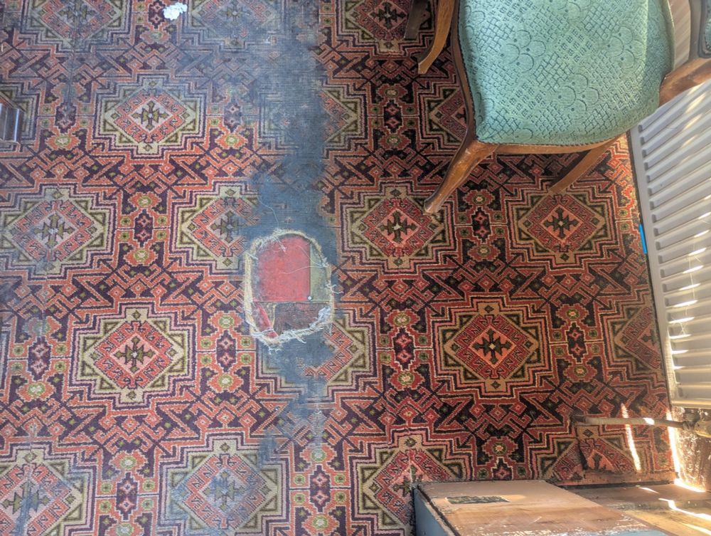 a picture of some patterned pub carpet. on the left half of the picture it is badly faded and marked, scuffed into only grey marks in some places and with a substantial hole through not just the carpet but the floorboards to the tiles below. on right right is a vibrant red carpet with clear patterns, unblemished