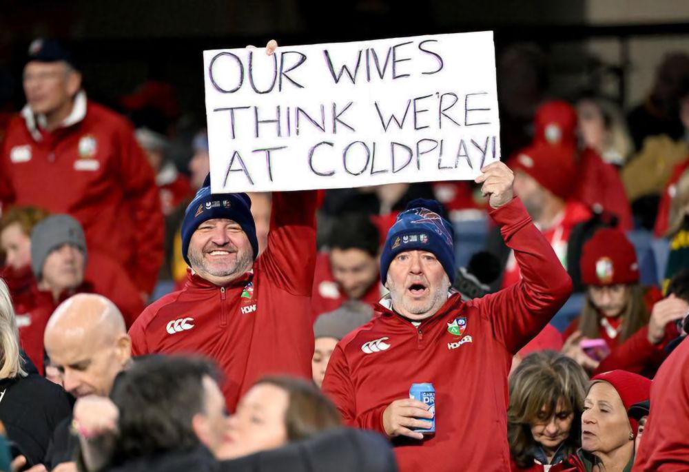 Two men at an event holding up a sign that reads "Our wives think we're at Coldplay" 