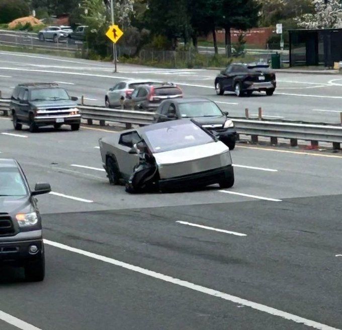 Photo of a cybertruck stopped in tbe middle of a busy highway. It's broken down, one of the tires is nearly off and twisted strangely. 