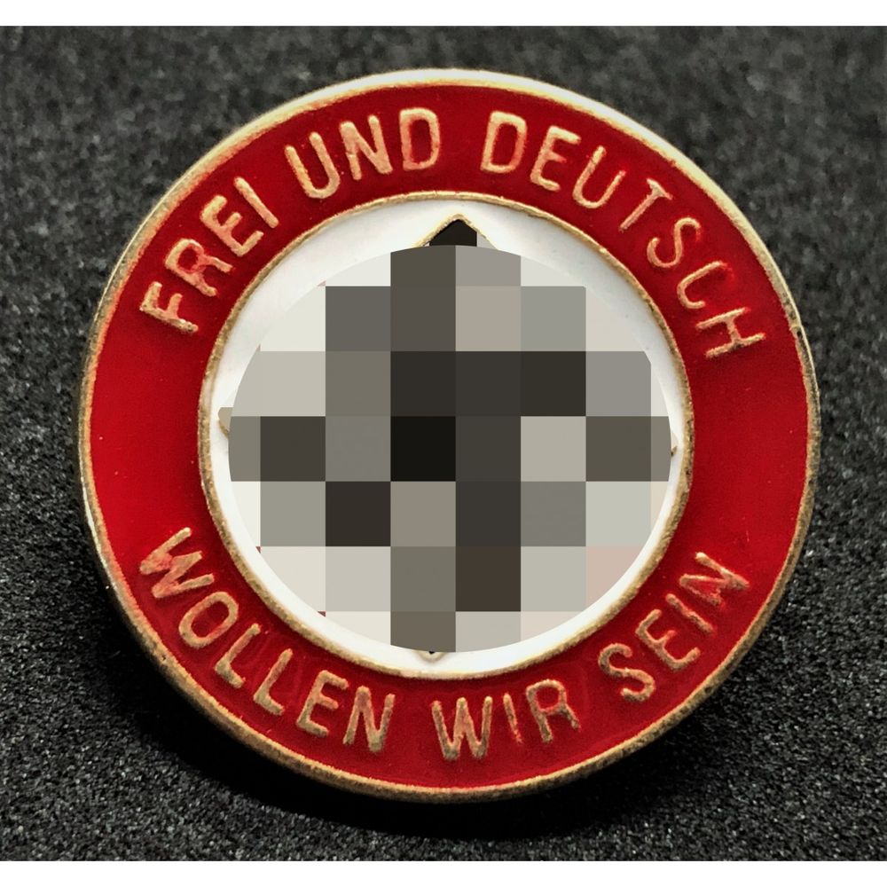 Metallabzeichen der Nazis mit der Aufschrift "FREI UND DEUTSCH WOLLEN WIR SEIN" am Rand und in der Mitte ein verpixeltes Hakenkreuz.