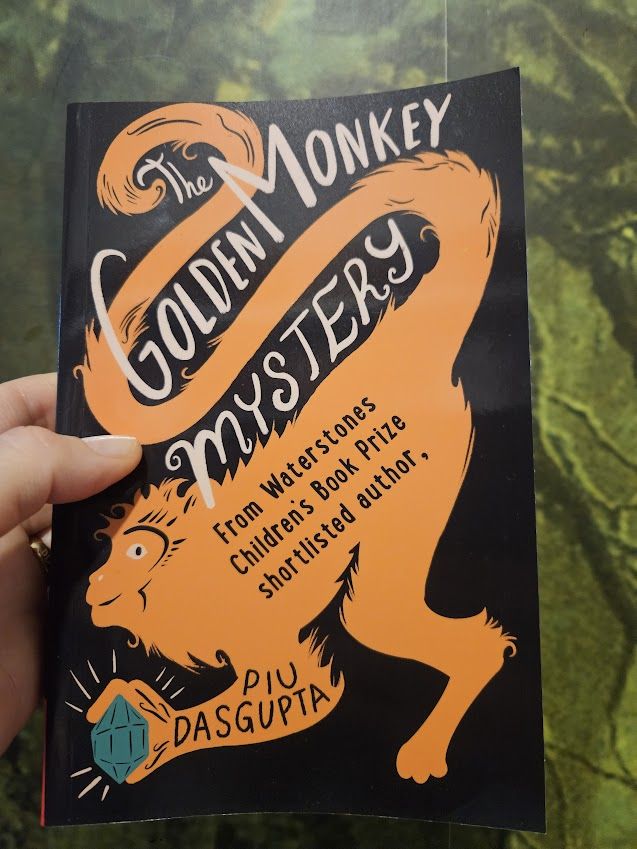 My hand holding up a book. The background of the cover is black and on it is a stylised orange monkey holding a greenish blueish jewel. The white, hand-lettered title, curving around the monkey and its tail, reads The Golden Monkey Mystery. The author's name, Piu Dasgupta, is written on the monkey's arm in black. The background behind the book is my rainforest wallpaper which, accidentally, fits the theme of the book as it's set partly in a jungle in Bengal.