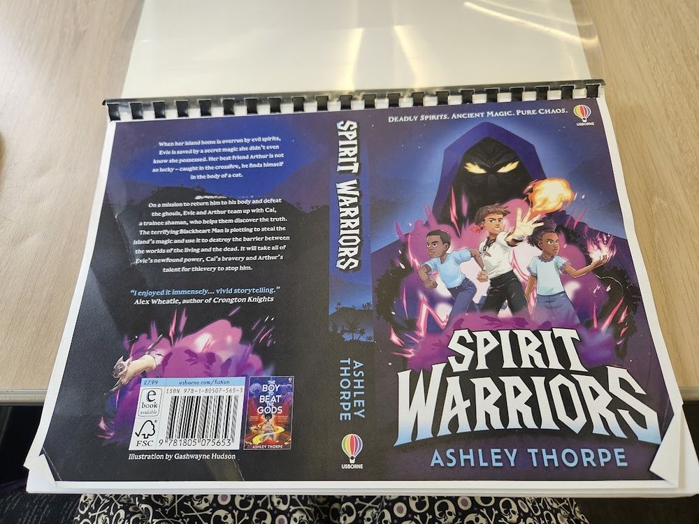 A proof of Spirit Warriors by Ashley Thorpe, laid out on a train table cos I was reading it on the train. The cover shows a mysterious shadowy hooded figure with glowing eyes. In front of him are three Black children, two are wielding magic. The far right girl hs a purplish magic, the central boy has fire magic. The other boy is running. The tag line is: DEADLY SPIRITS. ANCIENT MAGIC. PURE CHAOS. The overall colour vibe is purple.