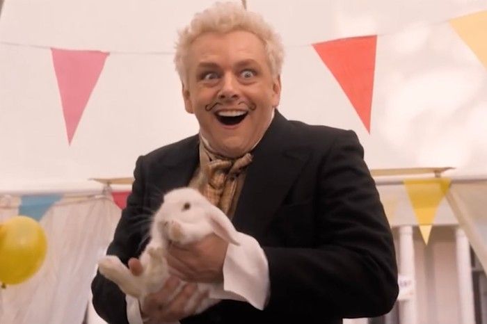 Aziraphale from season one of good omens holding a rabbit and looking ecstatic. He has a moustache drawn on and there is bunting behind him.