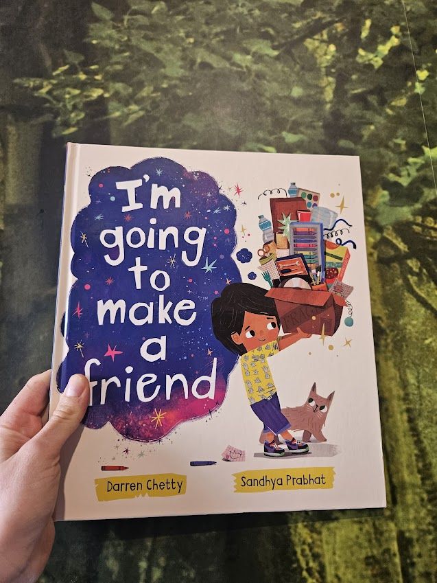 My hand holding a hardback picture book against wallpaper showing lush trees. The title is I'm going to make a friend and it's shown in a thought bubble full of stars and purple. A young boy with brown skin is carrying a big cardboard box of art equipment and random bits and pieces like spanners. The background of the cover is white and the author and illustrator names are in yellow flashes: Darren Chetty and Sandhya Prabhat. 