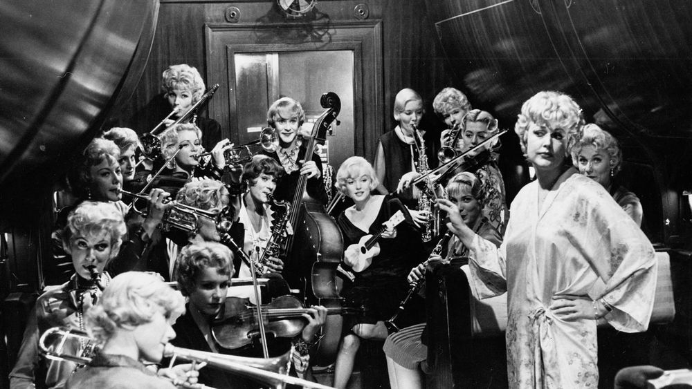 A black and white photo of the interior of a train car. A woman in a dressing gown is condiucting a disorganised looking band of white women, a lot of them with bottle blonde hair. If you look closely, two of them are Tony Curtis and Jack Lemmon dressed as women, and one is Marilyn Monroe with a tiny ukelele.
