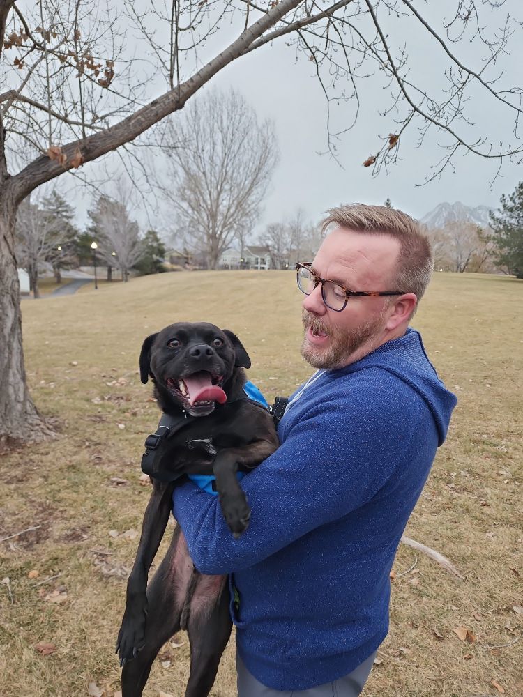 A lab/pug mix dog smiling at the camera with his tongue out, being held by a blonde man with a very short beard who is smiling at the dog. 
