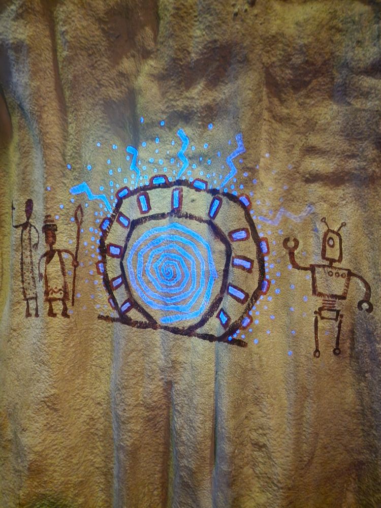 An artists rendition of The Stargate from picture one, designed to look like a cave painting with a glowing blue vortex in the middle. The gate is surrounded by three figures, somewhat stick-figured representations of an android style robot , one that is a shorter and more rounded robot, and one that appears like either a tall alien similar to the "Grays" or it may also be a robotic figure. I can not quite tell which. All are done in a style that suggests cave paintings somewhat like what the Ancestral Puebloans (sometimes inappropriately referred to as "Anasazi") would have drawn. 