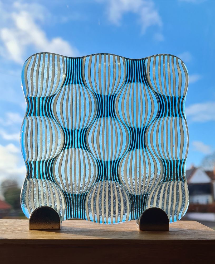 A square panel of glass in stripey blue and white where fusing has created a lens like circular pattern pushing the straight lines into curves.