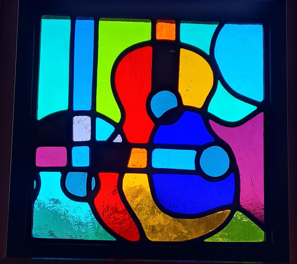 A square leaded stained glass panel showing brightly coloured overlapping guitars. The glass changes colour each time the shapes overlap.