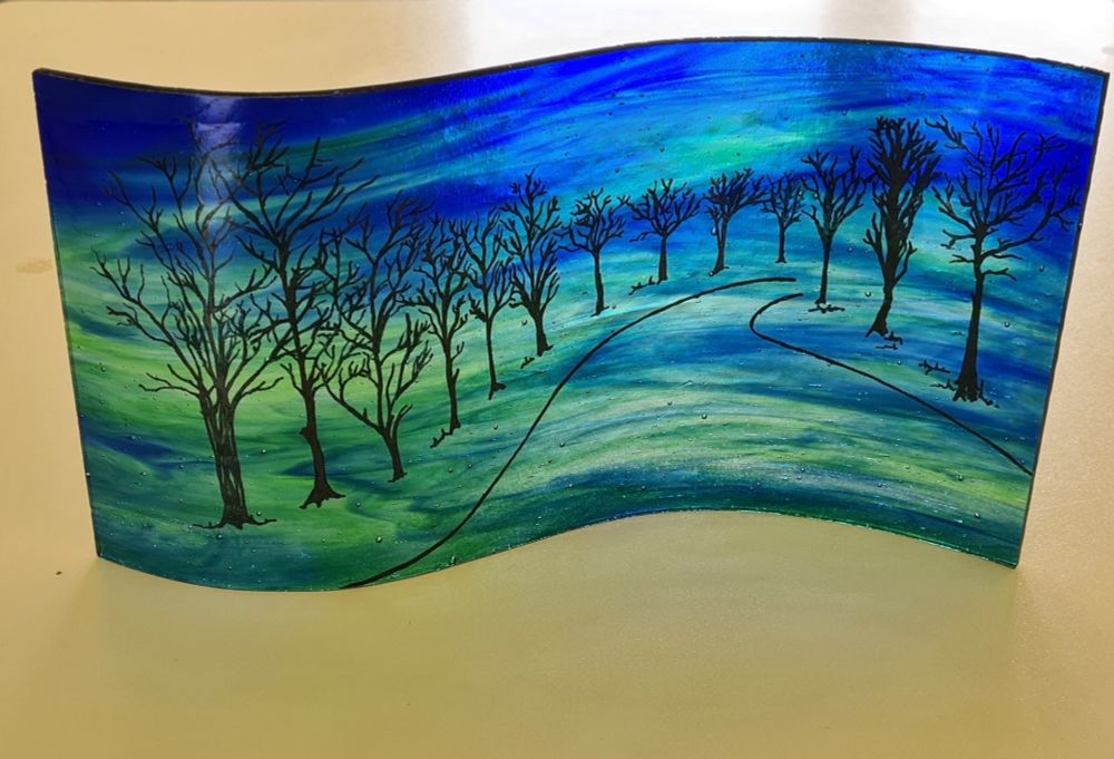 A blue and green wispy glass painted with a curved avenue of trees disappearing round a corner. This panel was slumped in an S mould so it curves and stands up on it's own.