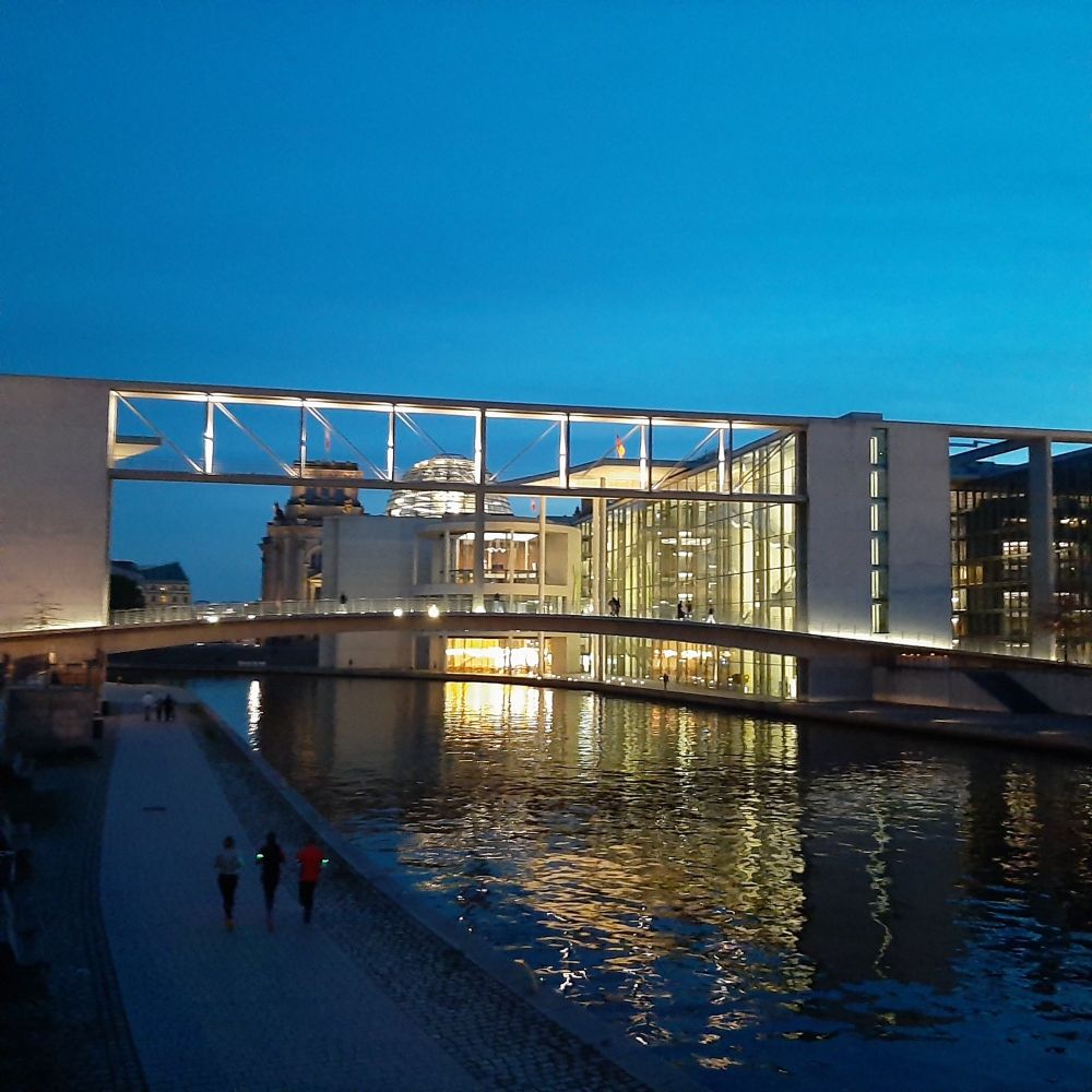 Das Bild zeigt das Reichstagsgebäude an See Spree in der Dämmerung. Himmel und Wasser sind sehr blau, das Innere der Gebäude hell erleuchtet.
