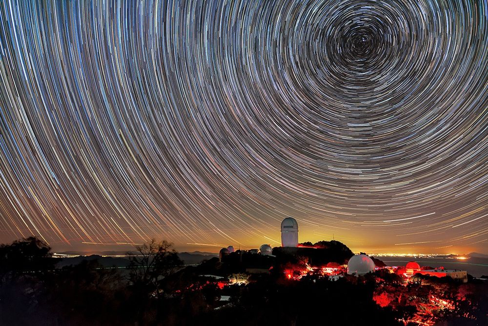 A long-exposure image captures circular star trails above telescope domes illuminated in red light on a dark mountain. A golden glow on the horizon marks distant city lights.
