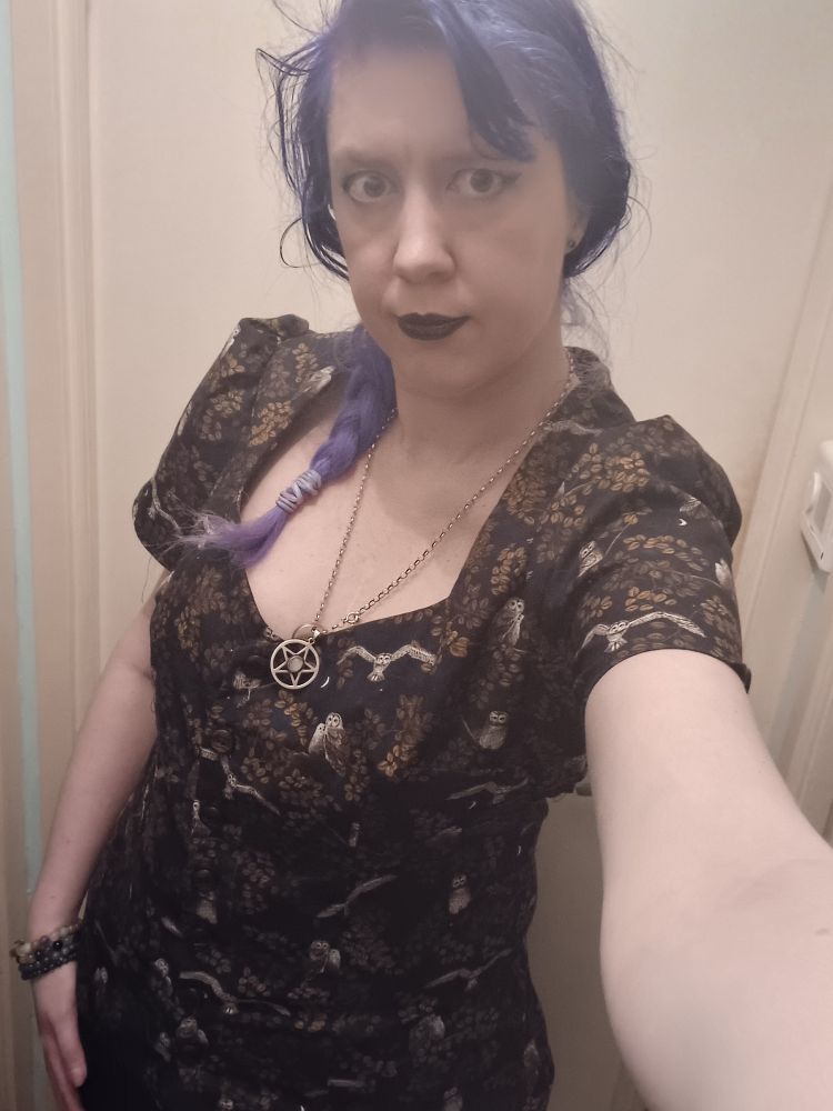 Photo of Jen, a white woman with green eyes, purple hair in a single braid woth purple lipstick, black eyeliner, purple and brown eyeshadow wearing a 50s style blouse with poofy short sleeves made out of a 100% cotton fabric with owl and leaves print on it that she sewed herself. A silver pentagram necklace is also visible