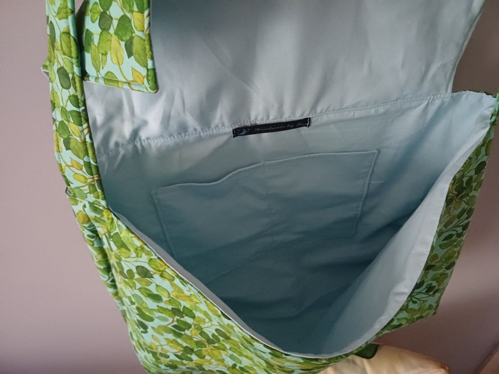 Inside of a large satchel bag made from 100% cotton green leaves on a blue background fabric & lined with 100% cotton light blue fabric. It has pockets inside & I made the magnetic buckle snaps on the front out of light blue faux leather with an iridescent rainbow finish

Black tag with a purple star and moon detail and purple writing reads "Handmade by Jen"