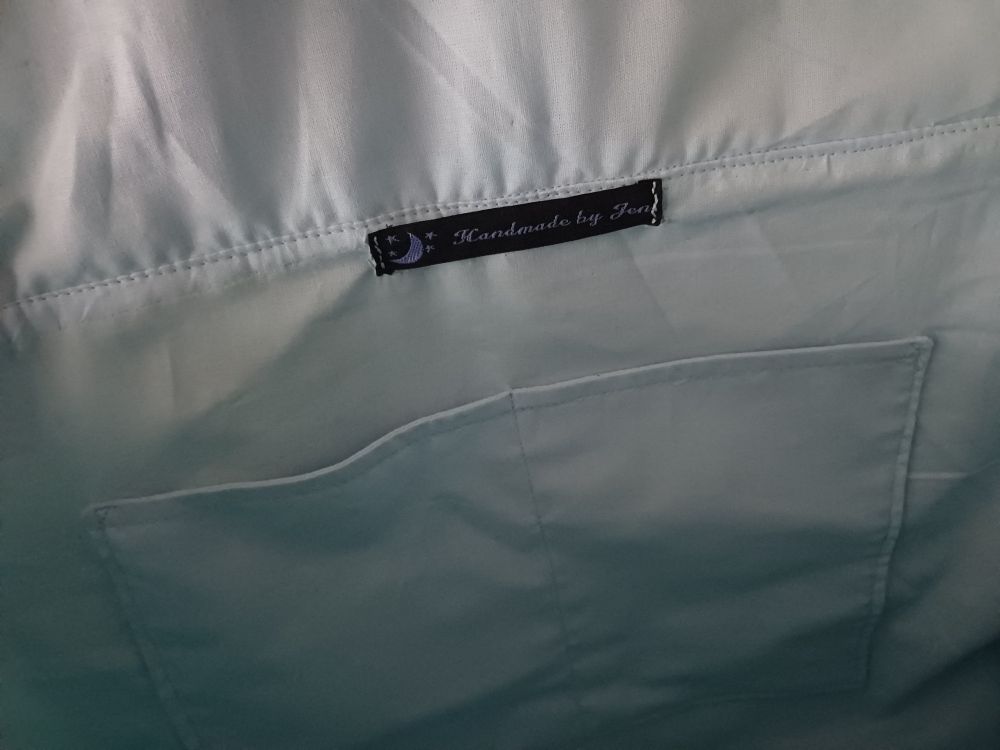 Inside of a large satchel bag made from 100% cotton green leaves on a blue background fabric & lined with 100% cotton light blue fabric. It has pockets inside & I made the magnetic buckle snaps on the front out of light blue faux leather with an iridescent rainbow finish

Black tag with a purple star and moon detail and purple writing reads "Handmade by Jen"