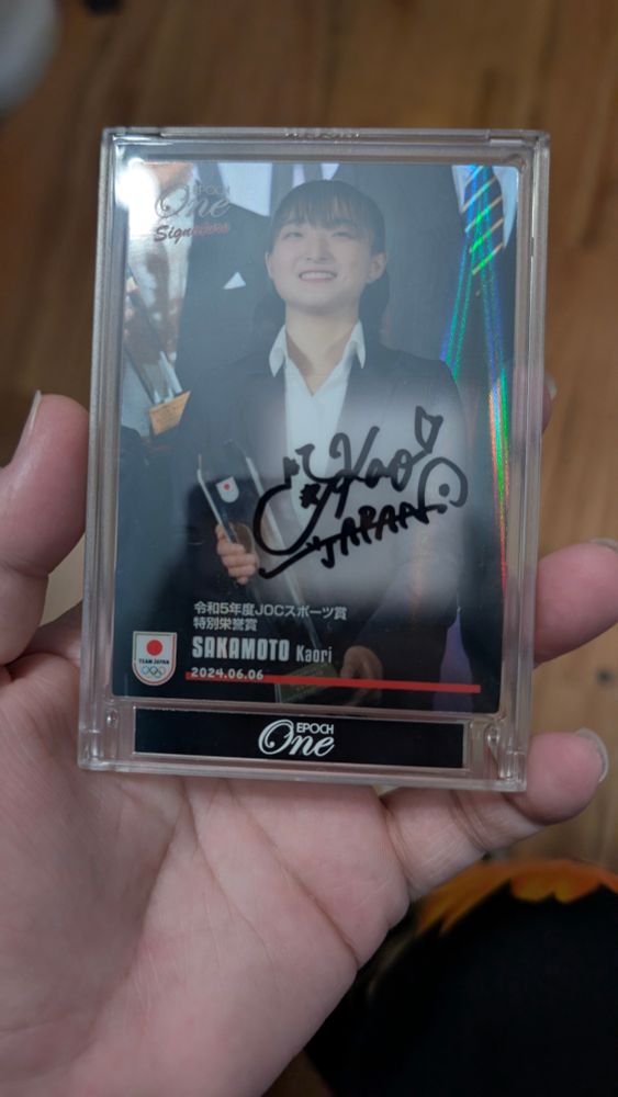 A trading card sealed in an acrylic case, featuring both the photo and autograph of Japanese figure skater Kaori Sakamoto. Kaori is wearing a suit and holding a glass trophy, smiling big for the camera. 
