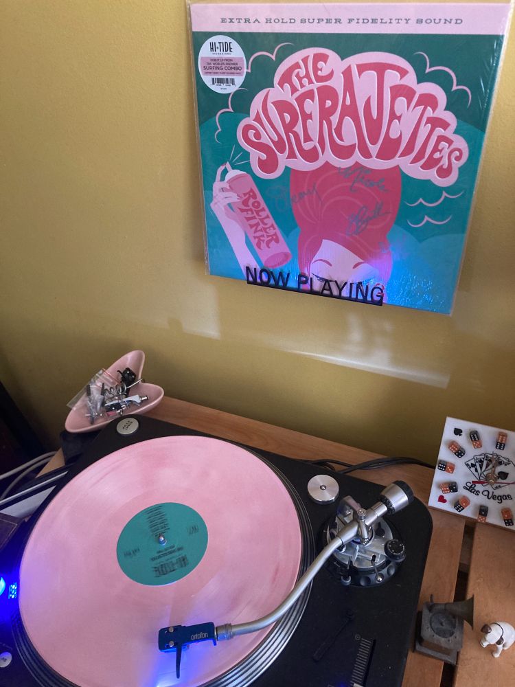 The Surfrajettes' debut album Roller Fink on pink vinyl. Record on a turntable, album cover (autographed by all four of the 2022 Surfrajettes) on a "NOW PLAYING" wall display