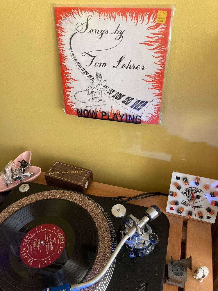 Ten-inch version of Songs by Tom Lehrer - record on a turntable exposing a 12-inch cork mat. Album artwork on a "NOW PLAYING" wall decoration above the turntable. 