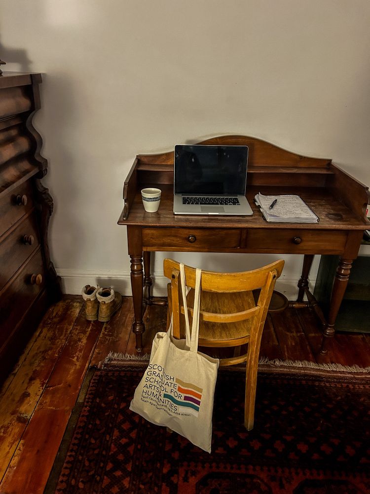 Old writing desk with a laptop, paper and cup of tea. A pair of boots are beside and a light coloured chair is in front, with a white canvas bag hung on the back. 