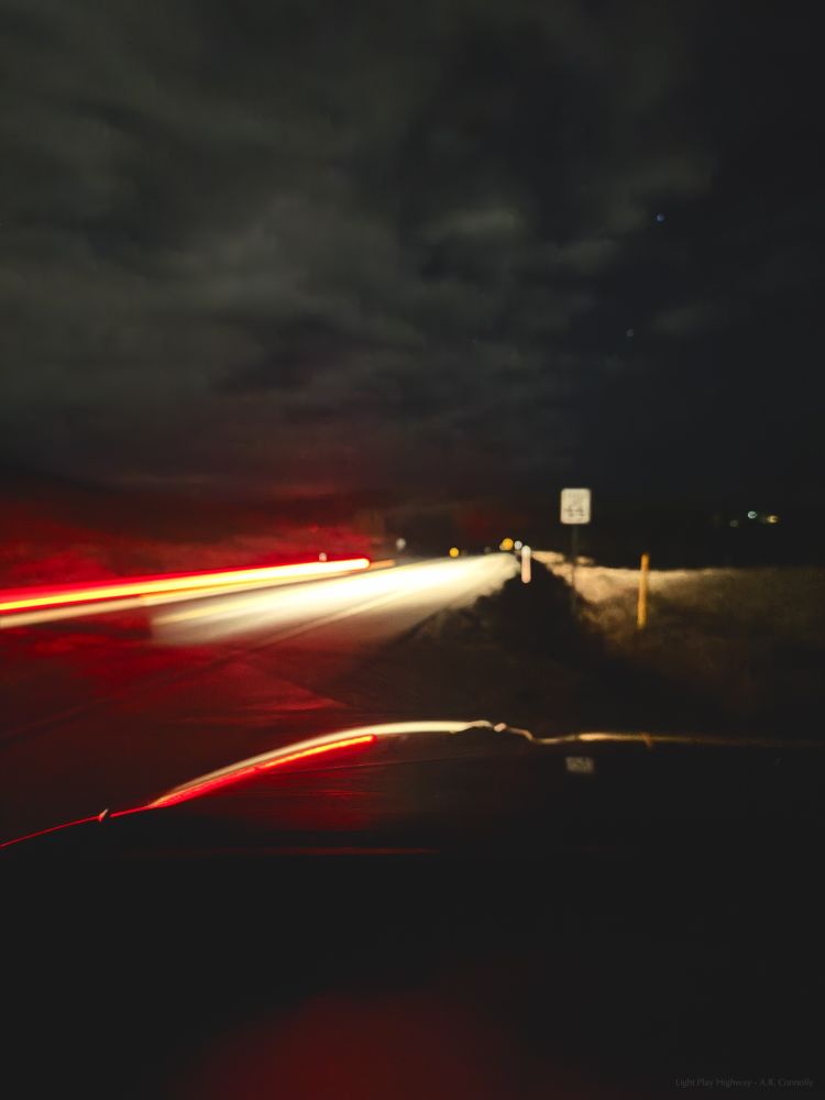 A photo of a two lane highway late at night. Most of the image is blurred, with semi formless clouds, a speed limit sign that's hard to read, and light trails of a passing car. The trails are reflected along the ground and the hood of the car where the photographer is sitting. 

"Light Play Highway - A.R. Connolly"