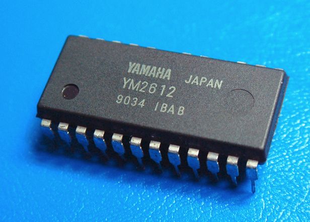 a picture of a Yamaha YM2612 soundchip - it is a 24-pin DIP package with 12 pins on either side, body itself is a matt dark gray with one glossy hole around the bottom left corner, and another less noticeable but larger matt hole towards the right side of the chip. there is text on it that says "YAMAHA Japan / YM2612 / 9034 IBAB"