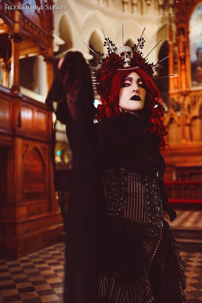 In the picture there is gorgeous red headed goth girl with black ruffle shirt, black and grey striped corset skirt, black mesh cape and a halo crown with roses and skull on it. In the background there is an altar of a church. the picture is taken in St. George Catherdral. 
